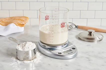 Glass sourdough crock on digital scale next to measuring cup full of flour